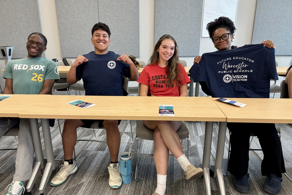 Four young people sit at a table, two of them holding up t-shirts.