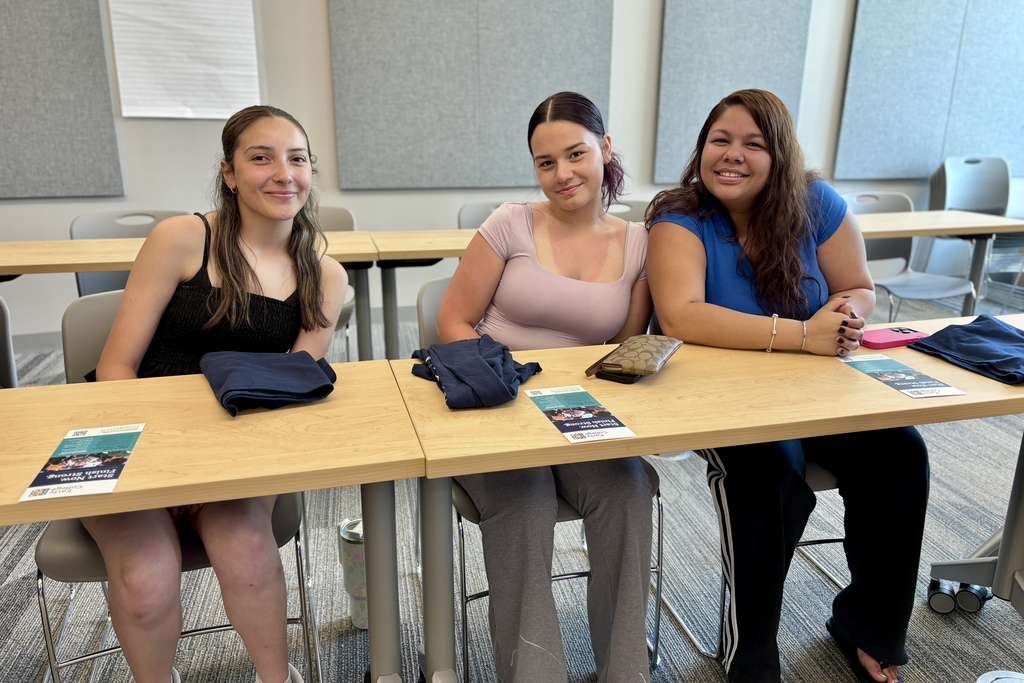 Three young women smile while sitting at a wooden table in a classroom setting.