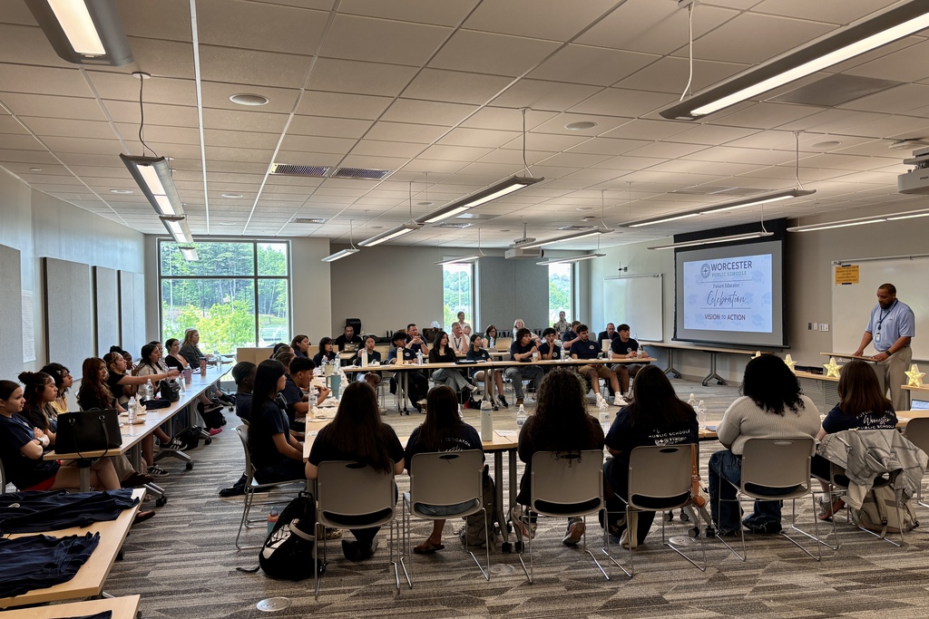 A group of students and adults are seated at tables in a conference room, listening to a presenter.