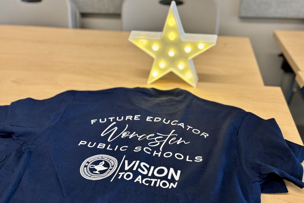 A navy blue t-shirt with white text reading 'FUTURE EDUCATOR Worcester PUBLIC SCHOOLS VISION TO ACTION' lies on a wooden table.