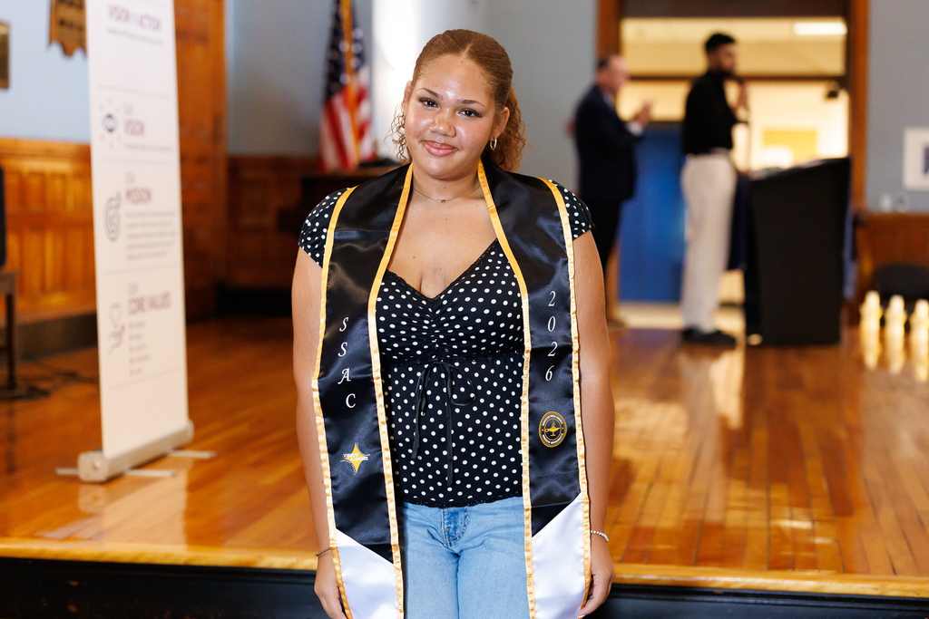 A student smiles while wearing a graduation sash.