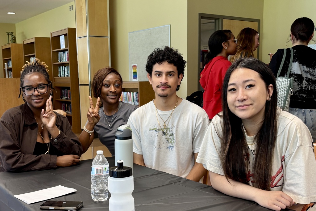 Four young people sit at a table in a room with bookshelves, smiling at the camera.