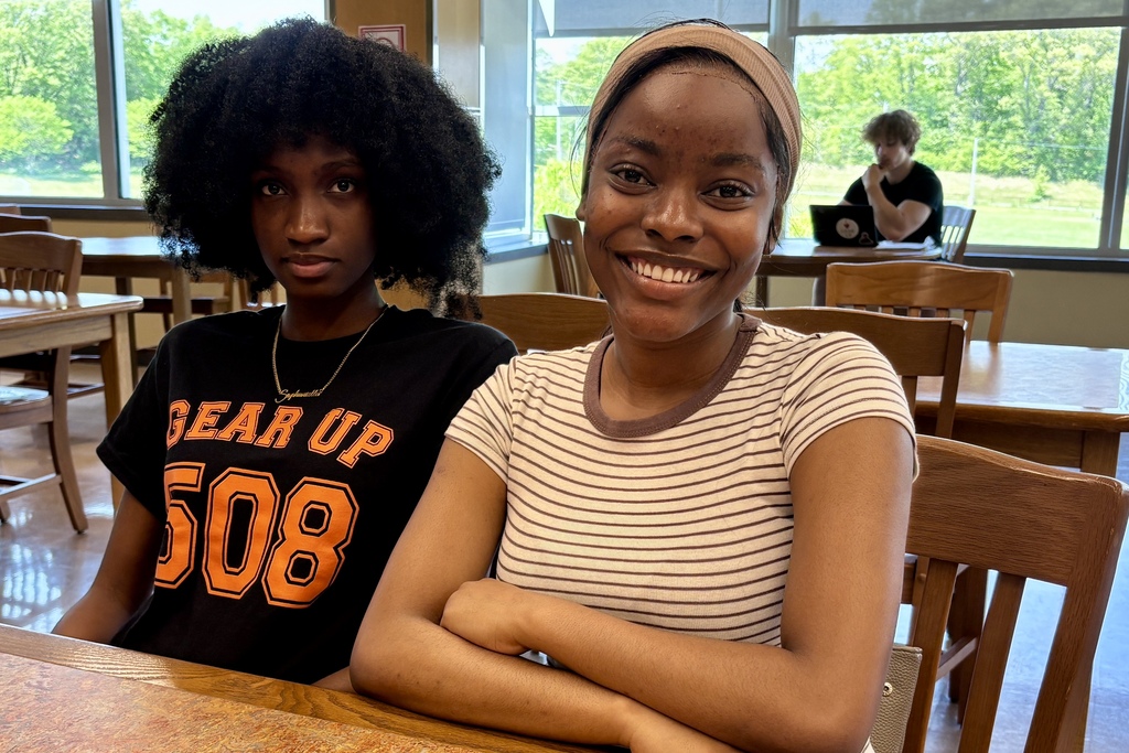 Two young women sit at a table in a room with large windows, one smiling and the other looking forward.
