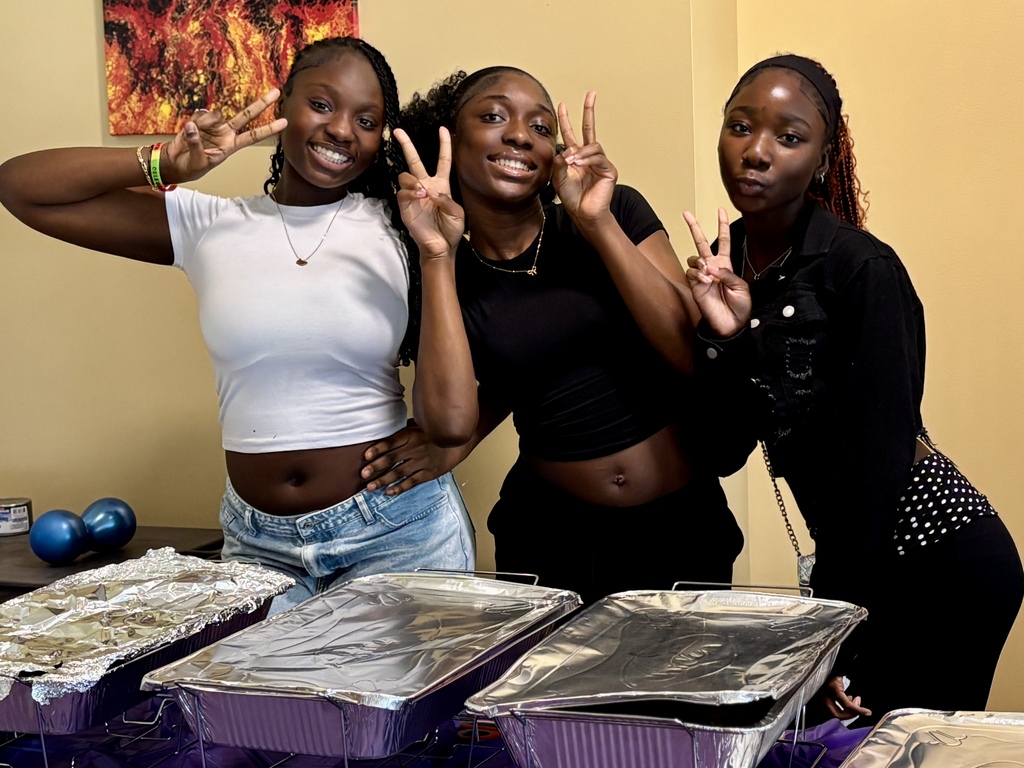 Three young women smile and pose with peace signs in front of foil-covered food pans.