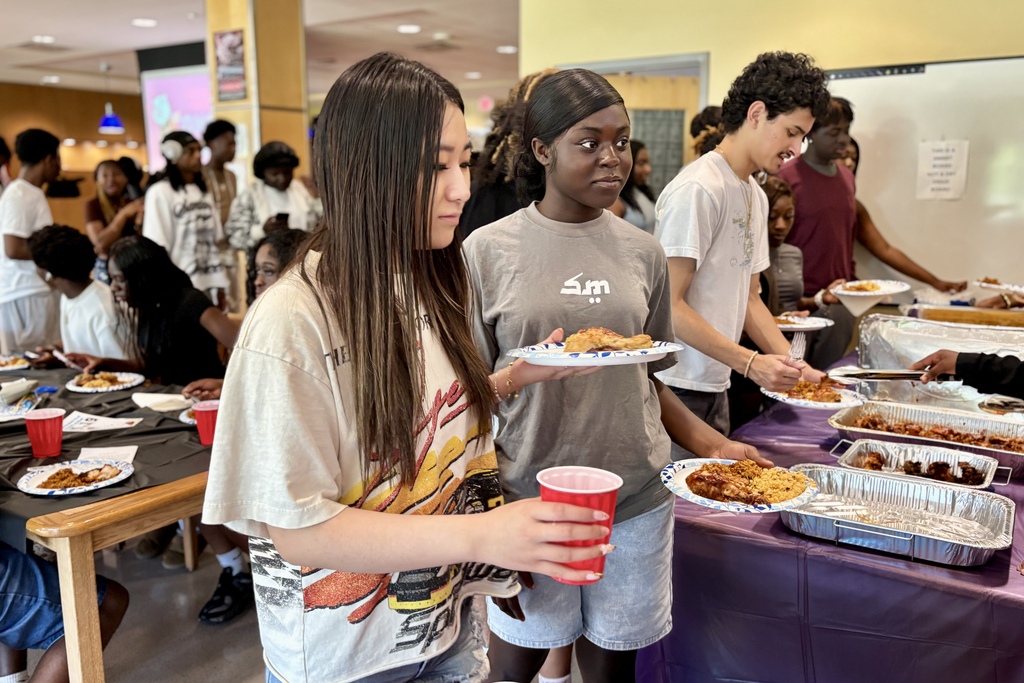 Young people gather around a table laden with food, serving themselves from foil trays.