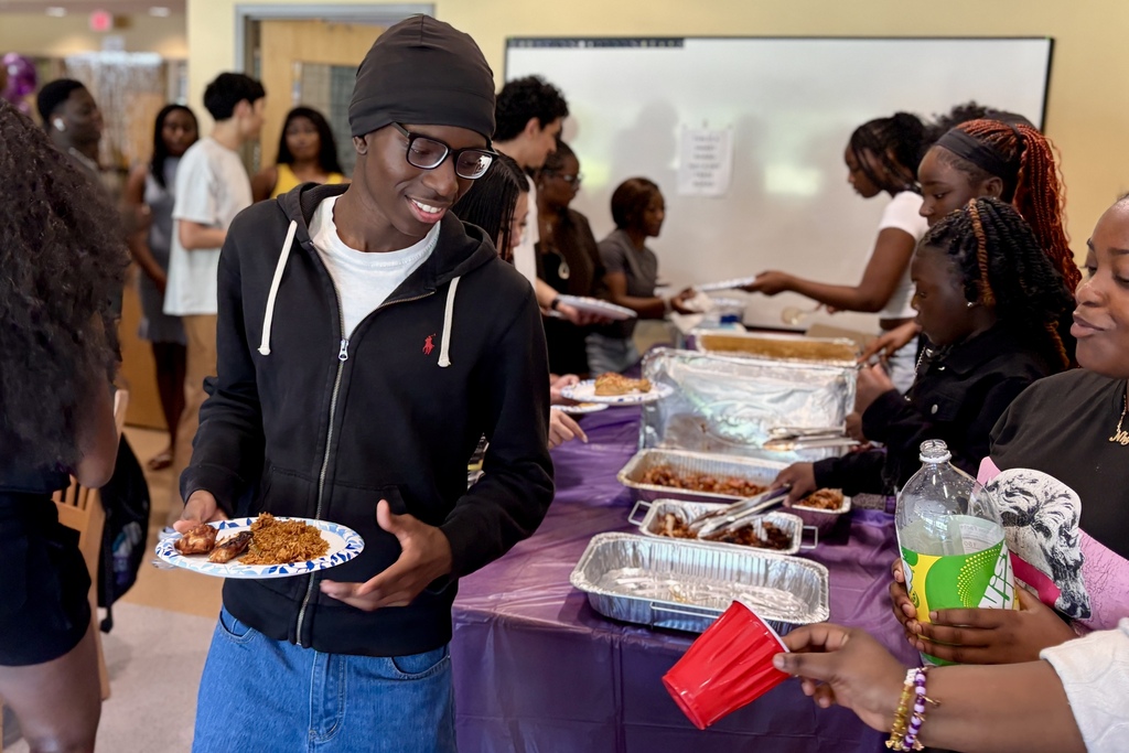 A young man with glasses and a black beanie smiles while holding a plate of food at a gathering.