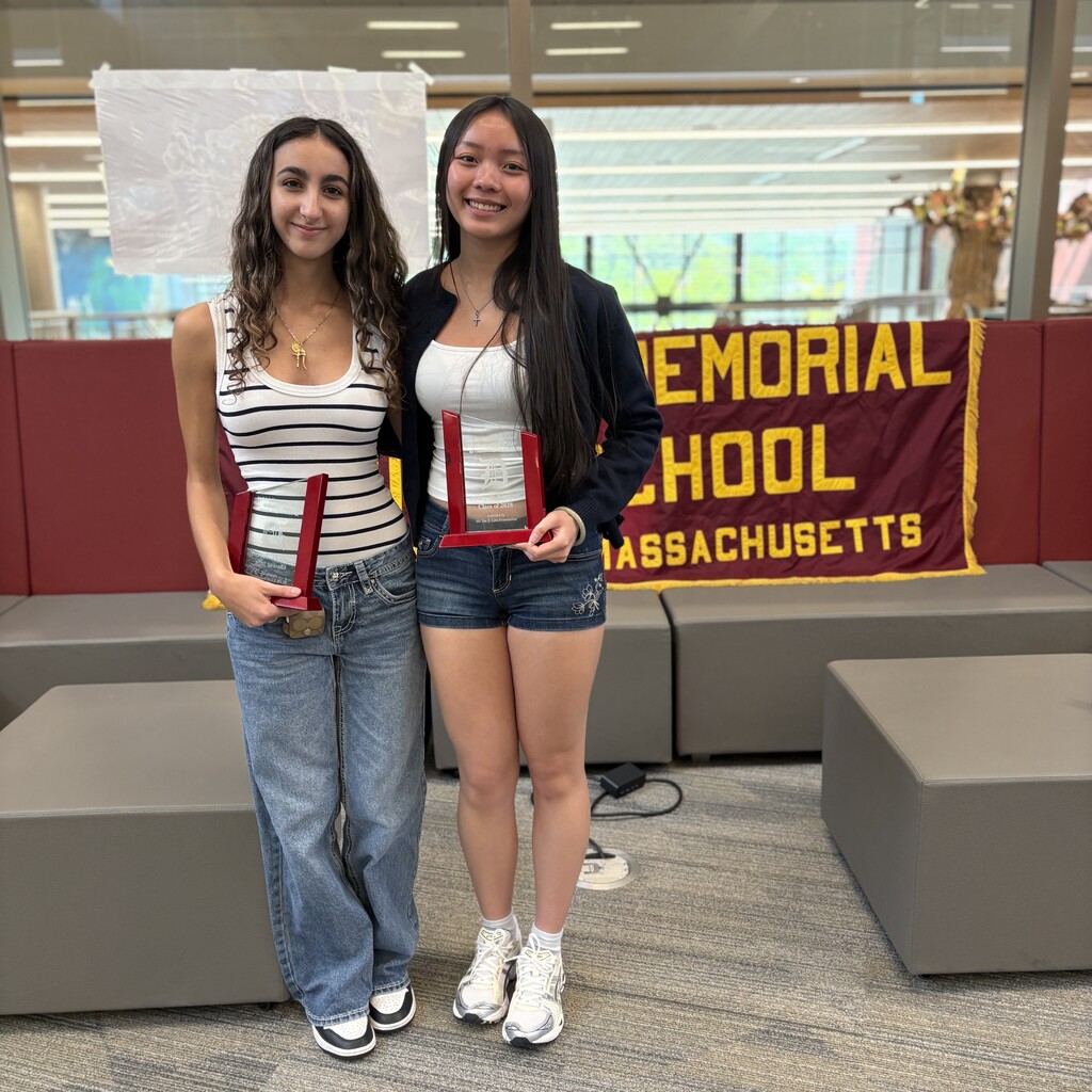 Two young women smile, each holding a red and glass award, in front of a school banner.