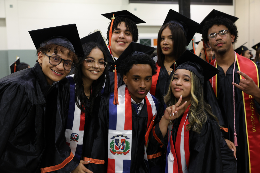 Seven students stand side-by-side posing and smiling while wearing graduation caps and gowns.