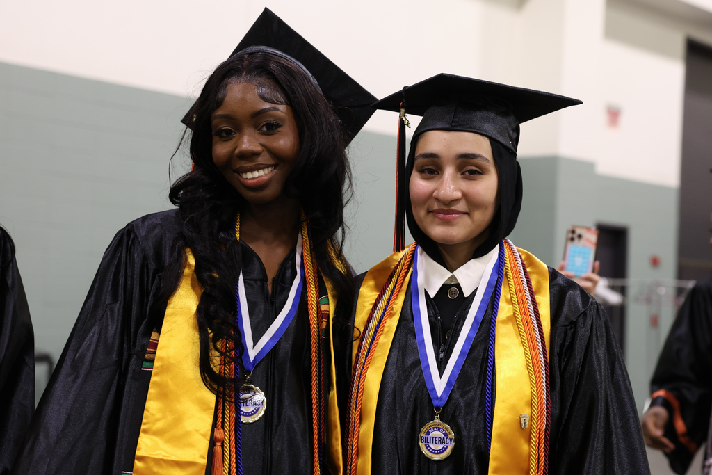 Two students stand side-by-side smiling while wearing their graduation cap and gown.