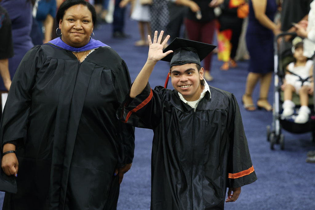 A student smiles and waves to the crowd while walking to stage during high school graduation.