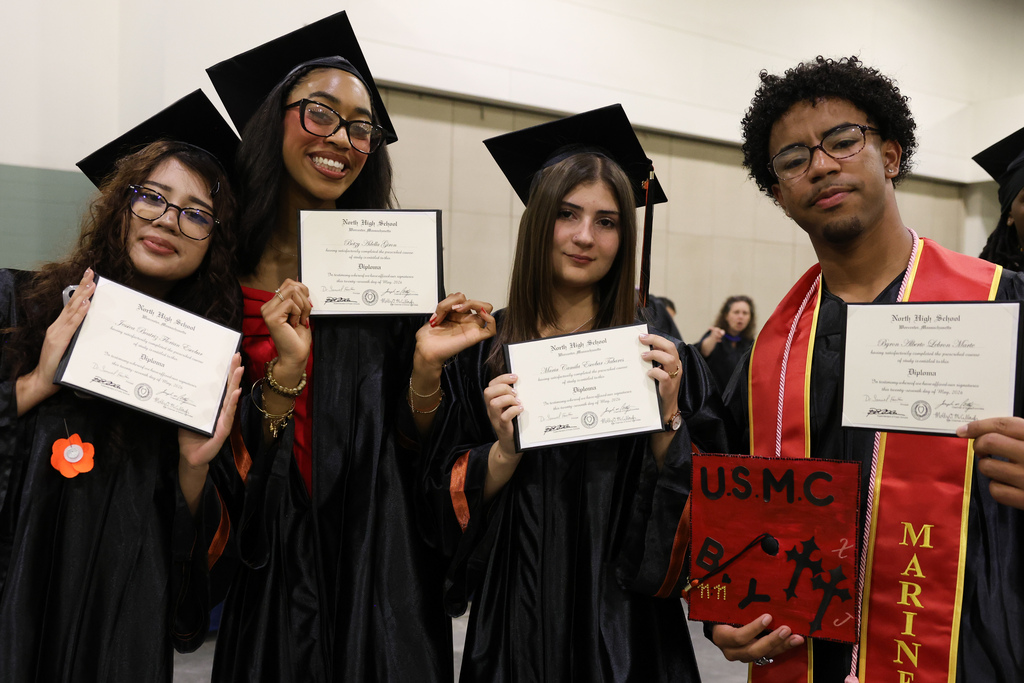 Four students stand side-by-side wearing their graduation gowns and holding up their high school diplomas.