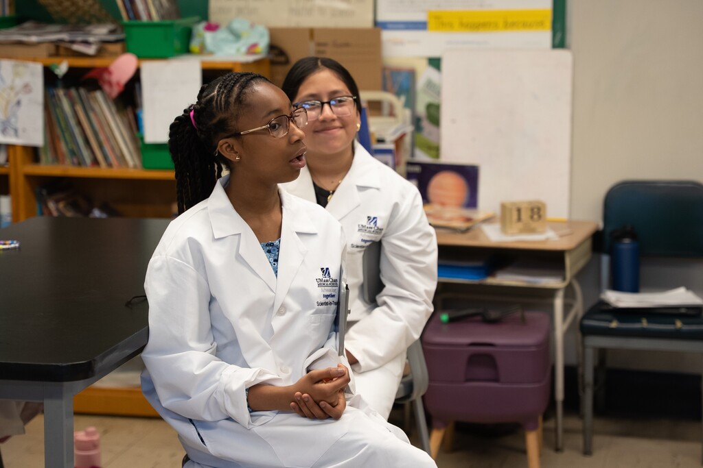 Two young students in white lab coats are in a classroom setting.