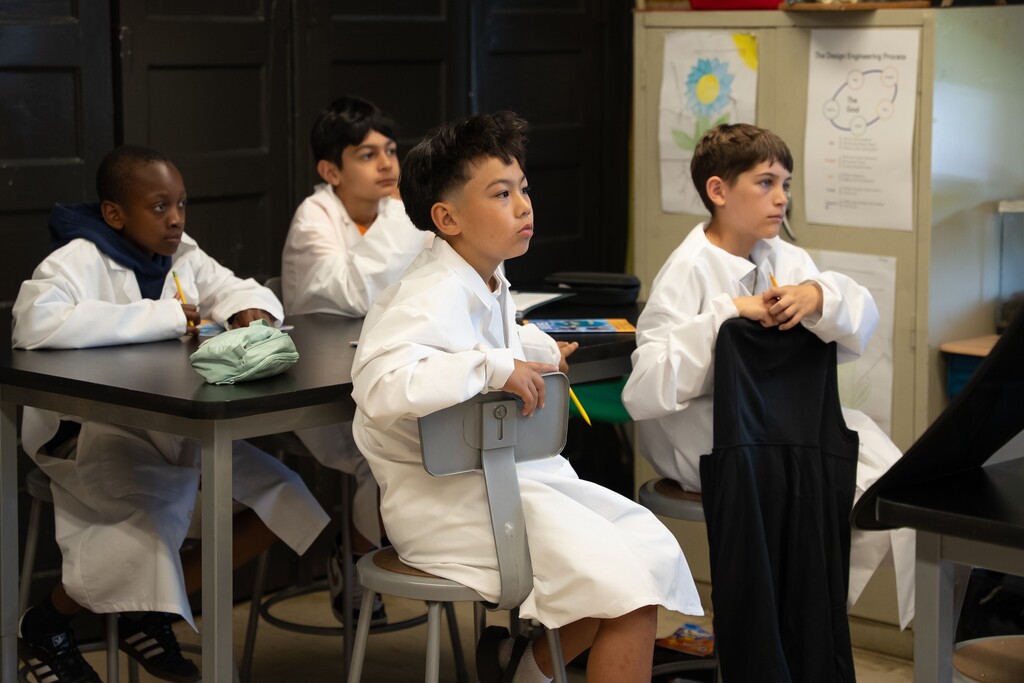 Two children in lab coats examine specimens in plastic containers with magnifying glasses and pencils.