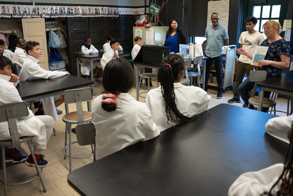 Students in white lab coats sit at desks in a classroom, listening to adults present.