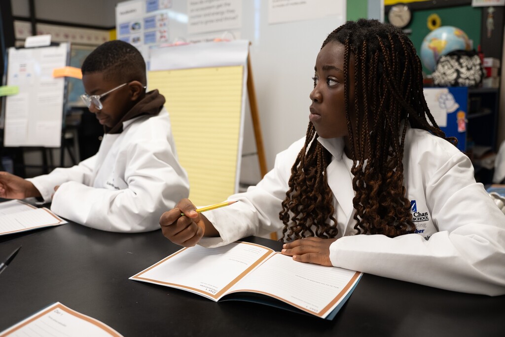Two students in lab coats are seated at a desk, engaged in a lesson.
