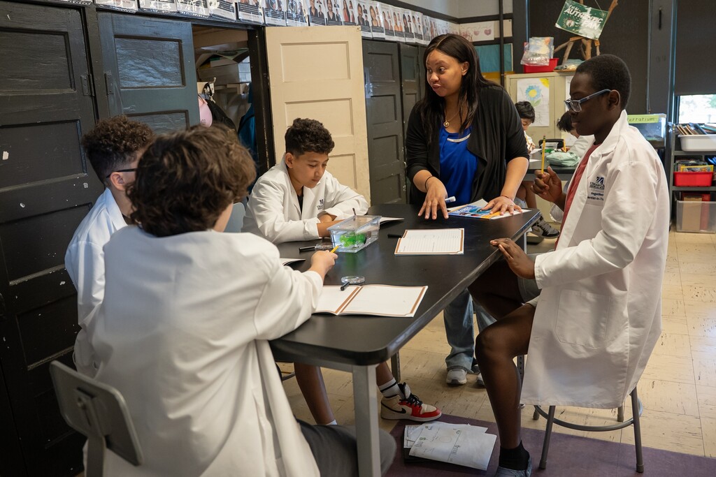 A teacher guides students in lab coats around a table with science materials.