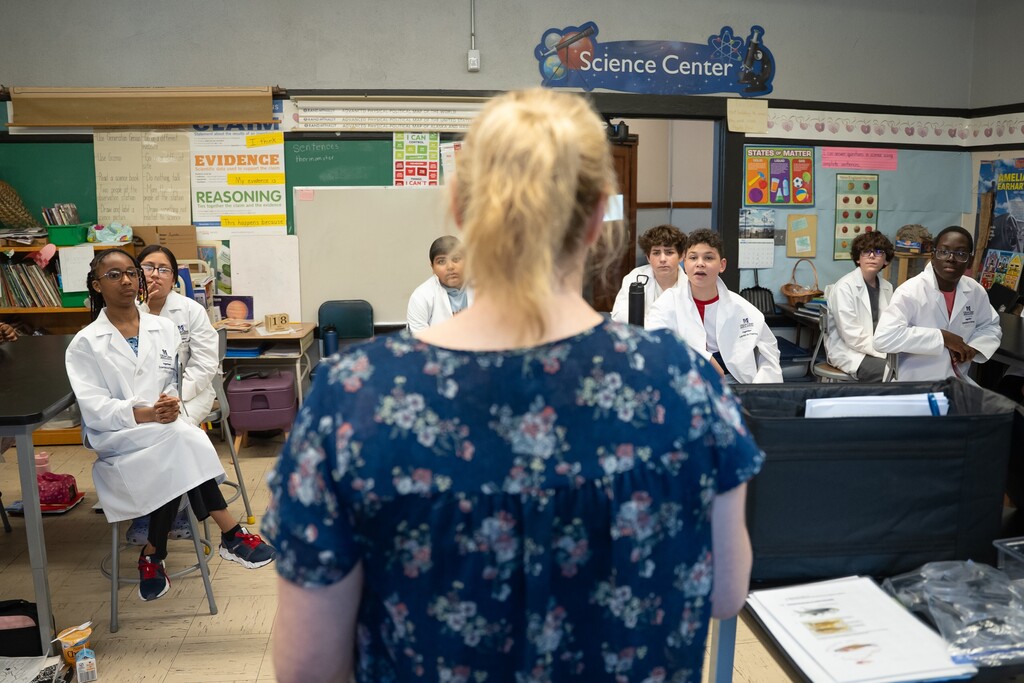 Students in lab coats listen attentively in a classroom with science-themed posters.