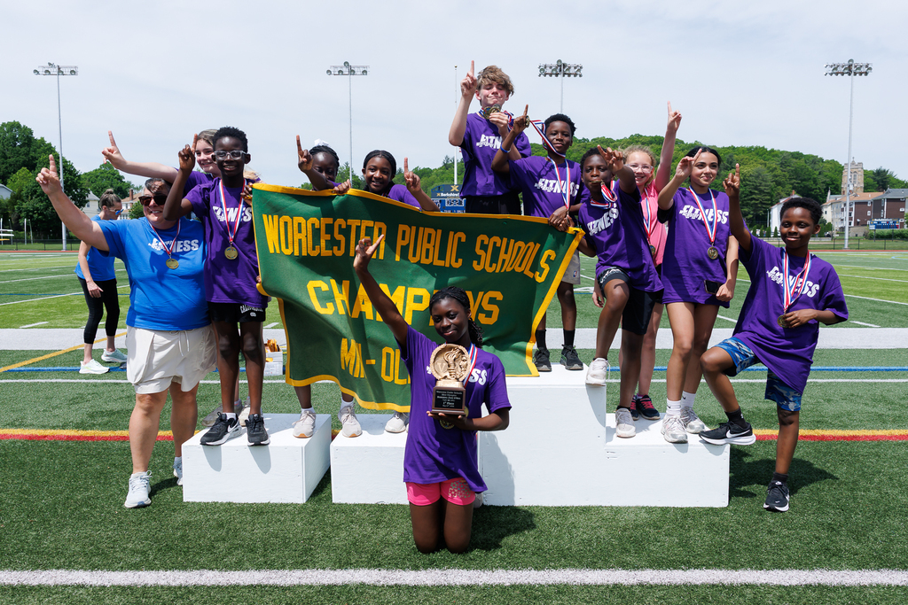 A group of students stand on a podium holding up the their pointer finger after winning a district-wide athletic competition.