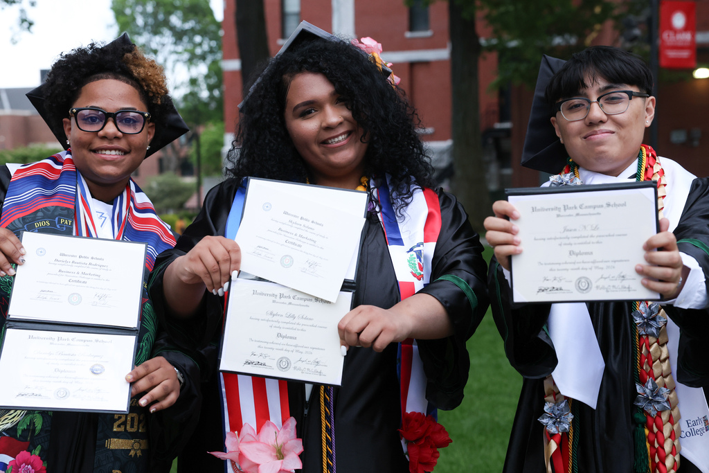 Three high school students wearing graduation caps and gowns smile while holding out their diplomas.