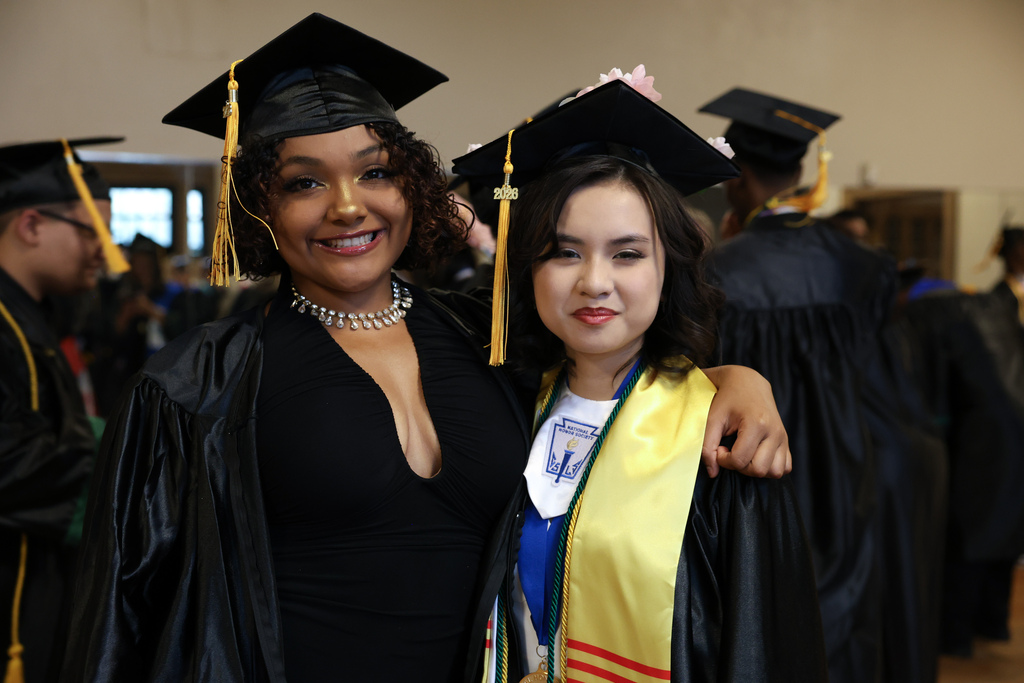 Two students wearing graduation caps and gowns smile while standing together.
