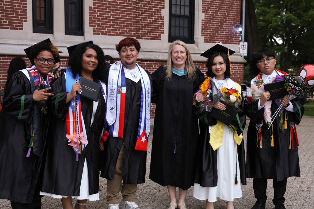 Five students in graduation caps and gowns hold their diploma while standing alongside their principal.