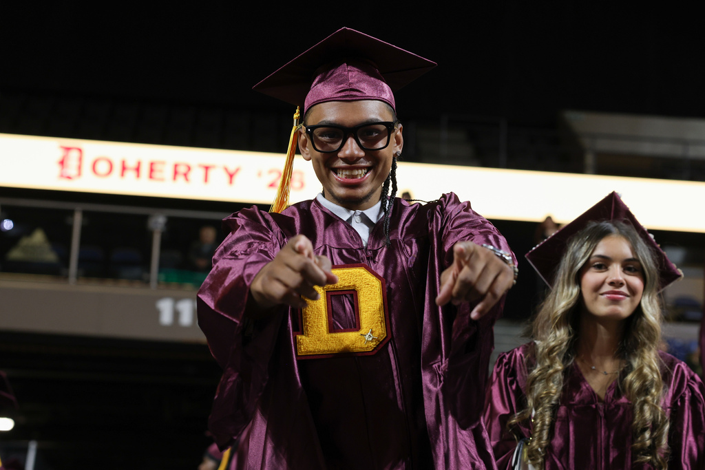 A student points to the camera while wearing a graduation cap and gown.