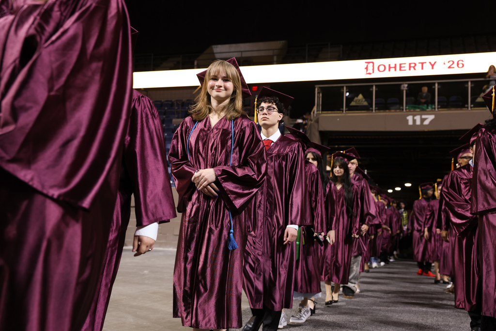 A student smiles while walking in line at their high school graduation, wearing a cap and gown alongside their classmates.