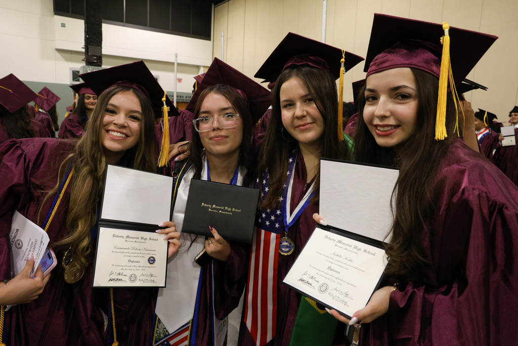 Four students hold up their diplomas while wearing graduation caps and gowns.