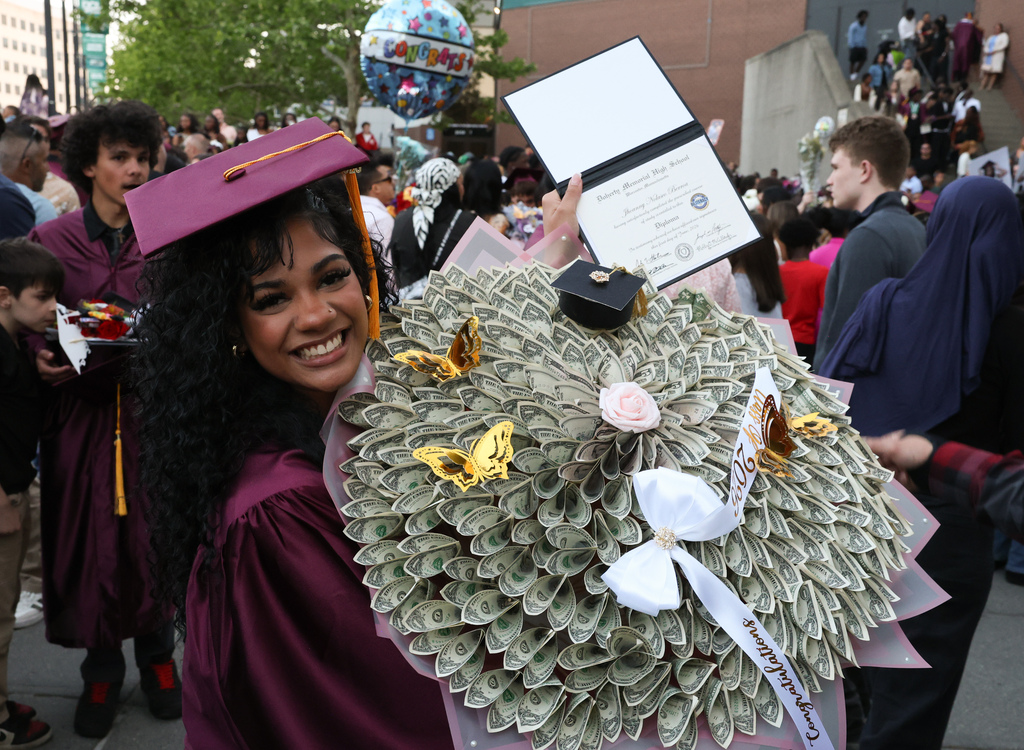 A student holds a bouquet of dollar bills in one hand and their diploma in the other following their high school graduation.