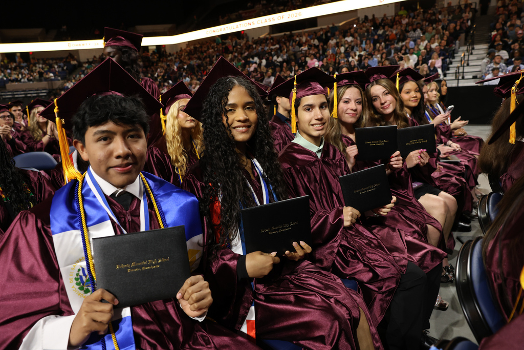Students sit in their chairs while wearing graduation caps and gowns and holding their diplomas.