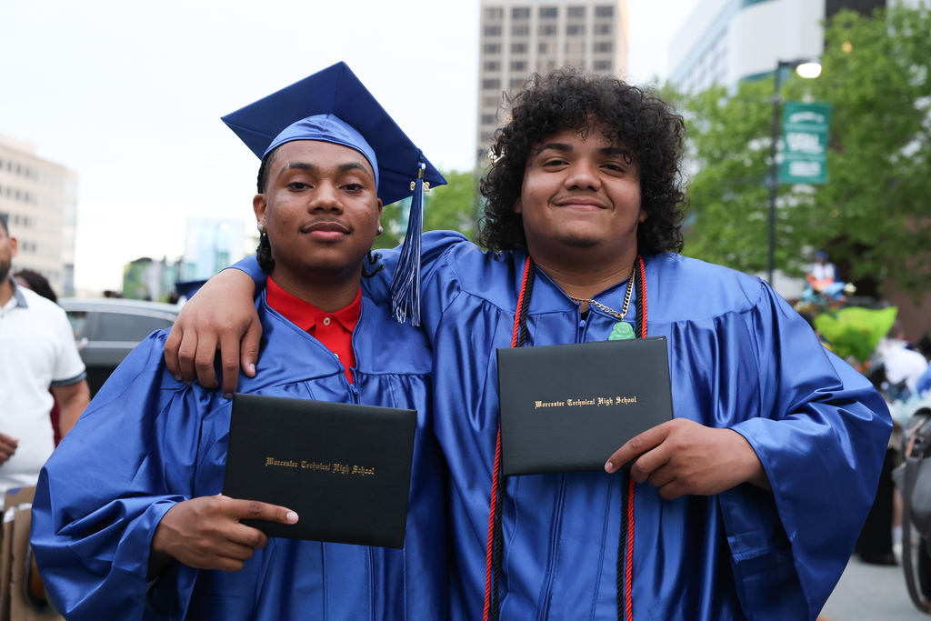 Two students wearing caps and gowns hold up their high school diplomas following graduation.