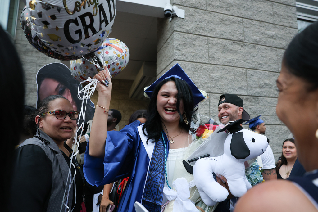 A student wearing a cap and gown holds up a balloon that says "congrats grad" while smiling wide.