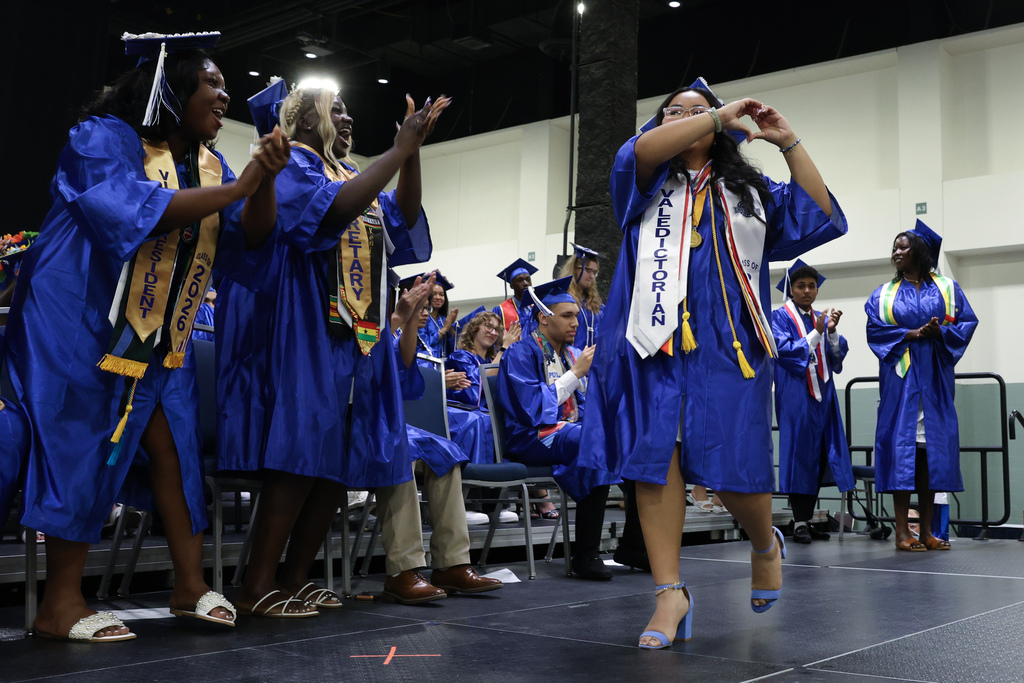 A high school student in a graduation cap and gown makes a heart symbol with their hands while other students cheer them on as they walk across stage at graduation.