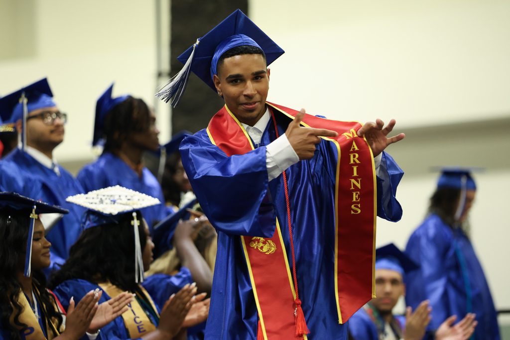 A student wearing a cap and gown points to a stoll that said "Marines" as they walk across stage at graduation.