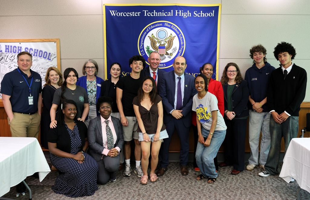 A group of students and adults pose for a photo in front of a banner for Worcester Technical High School.