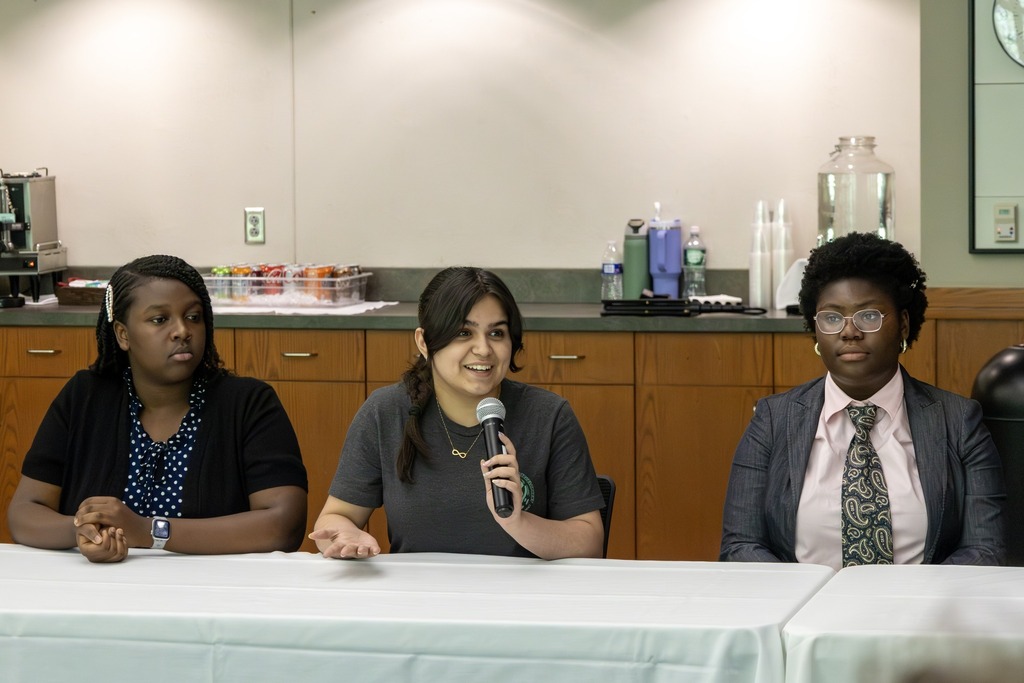 Three young women sit at a table, with one speaking into a microphone.