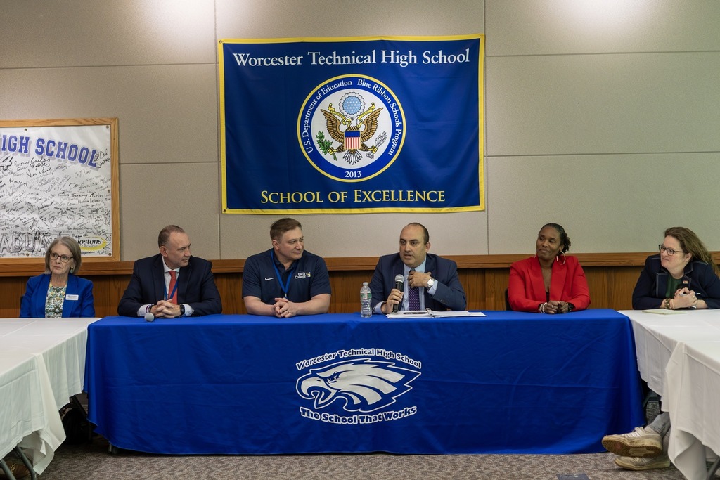 A panel of five individuals sits at a table in front of a banner for Worcester Technical High School, a School of Excellence.