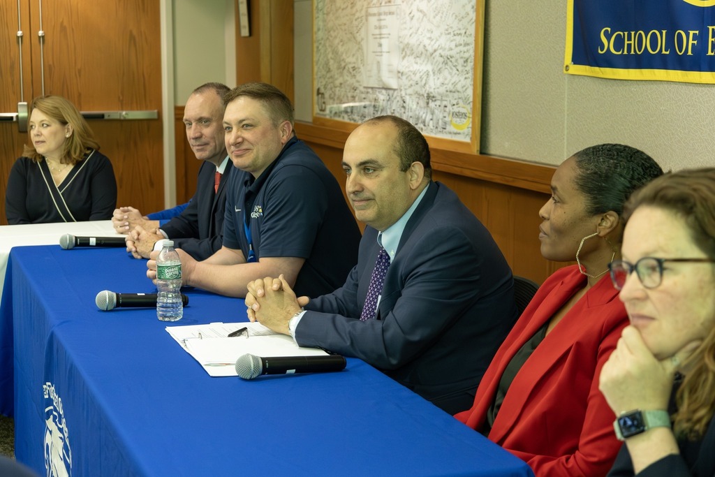 A panel of five individuals sits at a table covered with a blue cloth, with microphones and water bottles in front of them.