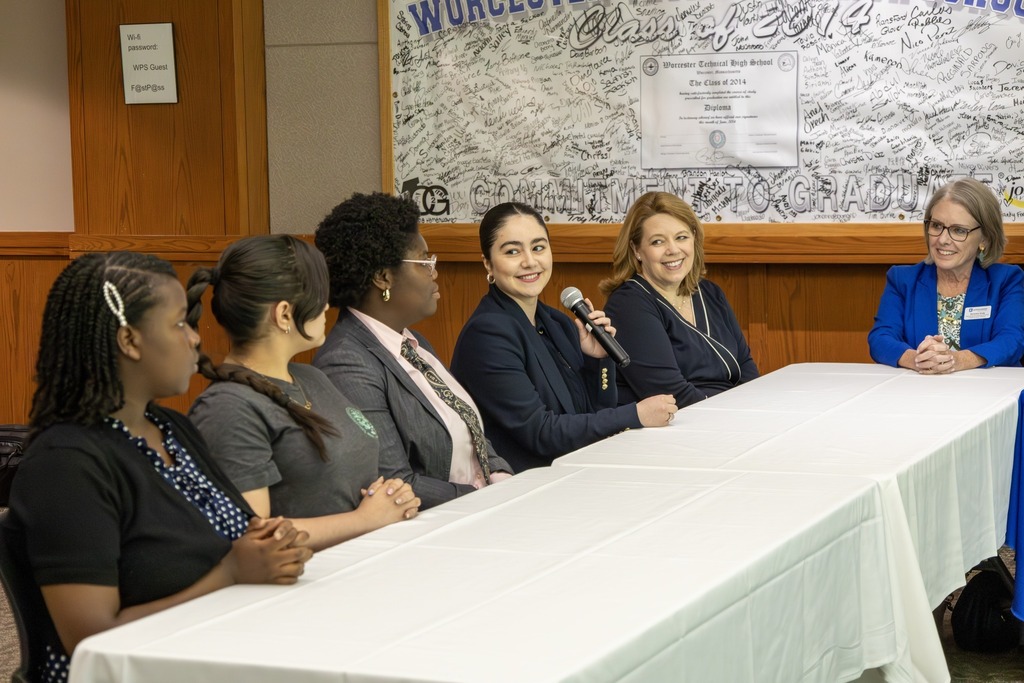 A panel of five women, including students and educators, sit at a long table in front of a wall covered in signatures and a diploma.