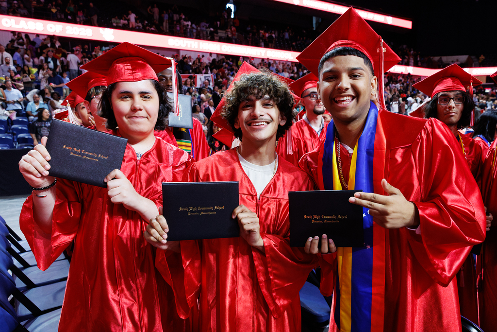 Three high school graduates hold up their diplomas while wearing caps and gowns.