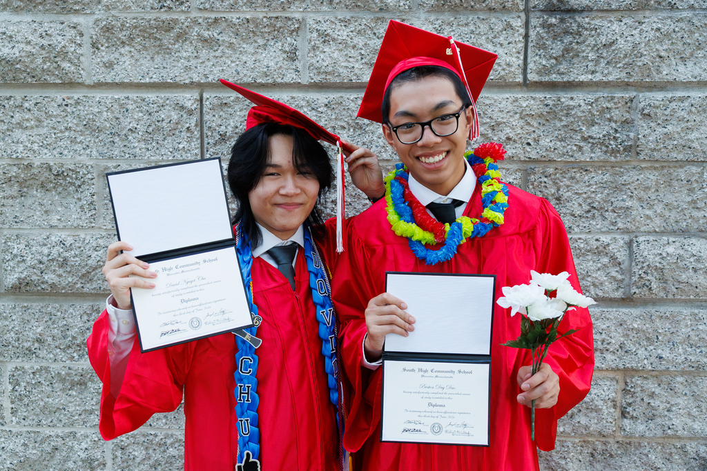 Two high school graduates wearing a cap and gown hold out their diplomas.
