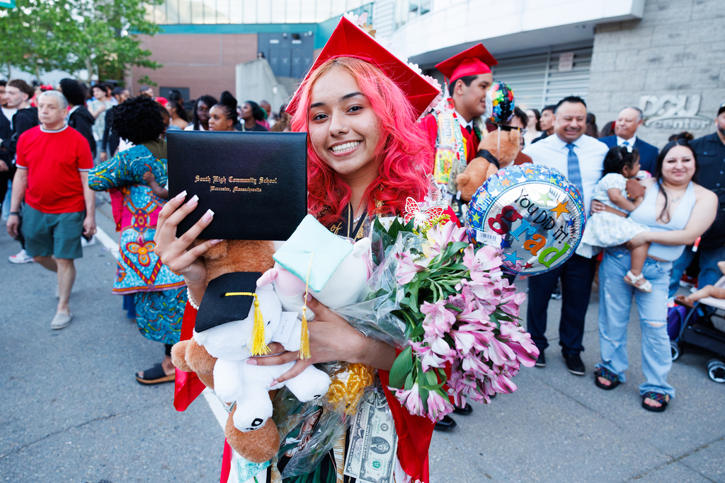 A student holds up their diploma while holding flowers, balloons and stuffed animals.