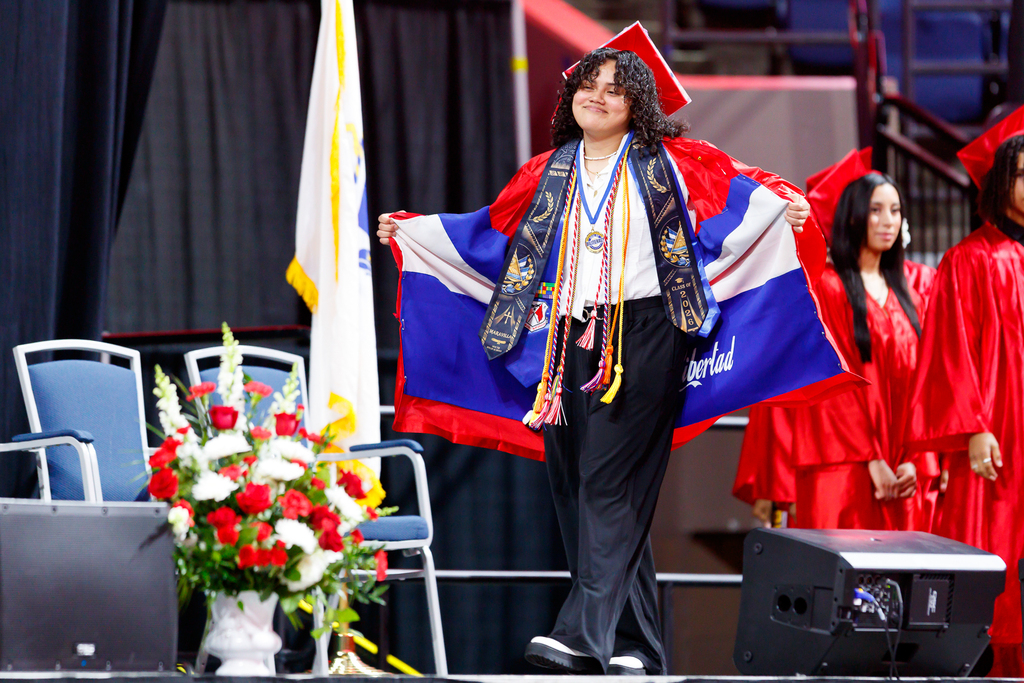 A student shows off a flag in their graduation robe lining as they walk across stage.