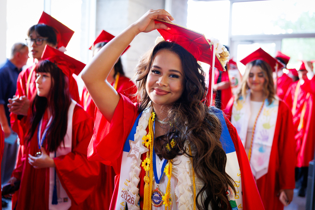 A student adjusts their graduation cap while walking through a well-lit hallway.