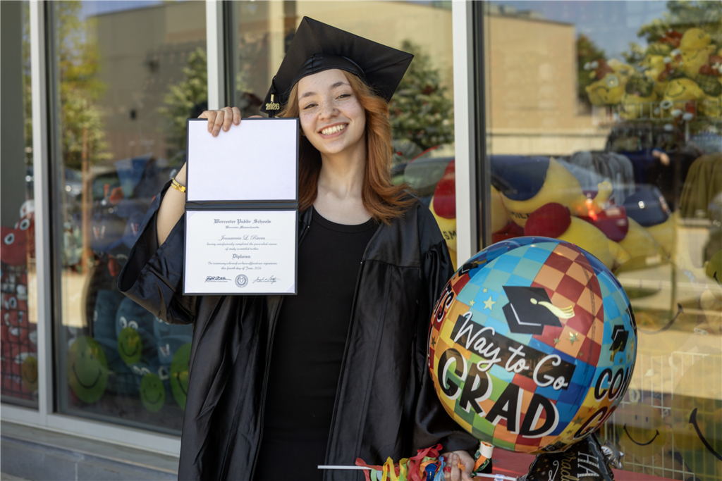 A smiling graduate in a cap and gown holds a diploma and a colorful "Way to Go GRAD" balloon.
