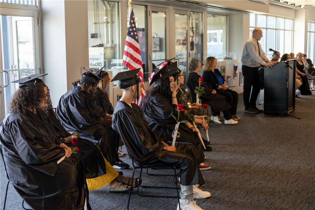 Graduates in black gowns and caps sit in chairs, holding red roses at a ceremony.