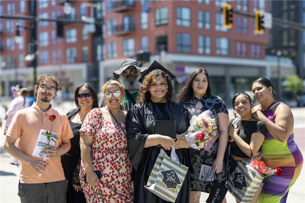 A group of smiling people, some in graduation attire, pose for a photo outdoors with buildings in the background.