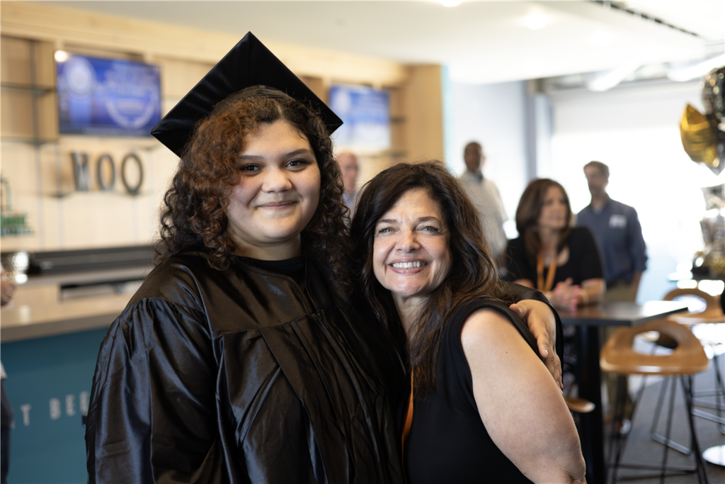 A graduate in a black cap and gown smiles with a woman in a black top.