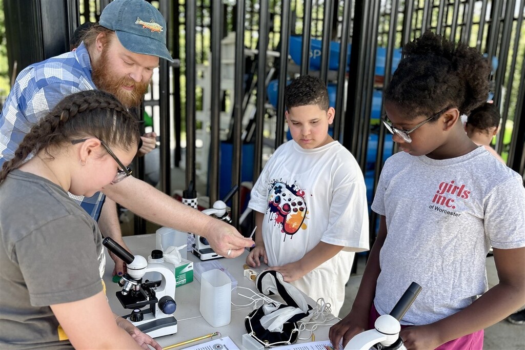 A group of children and an adult are gathered around a table with microscopes, engaged in a science activity.