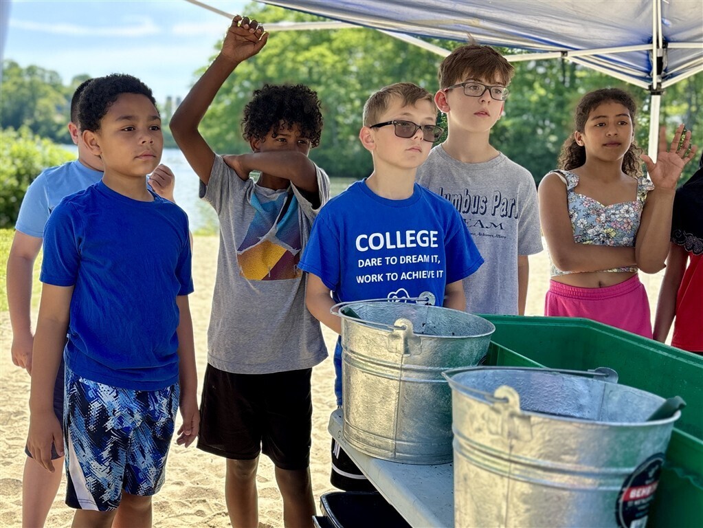 Children stand by a table with buckets, looking towards the water.